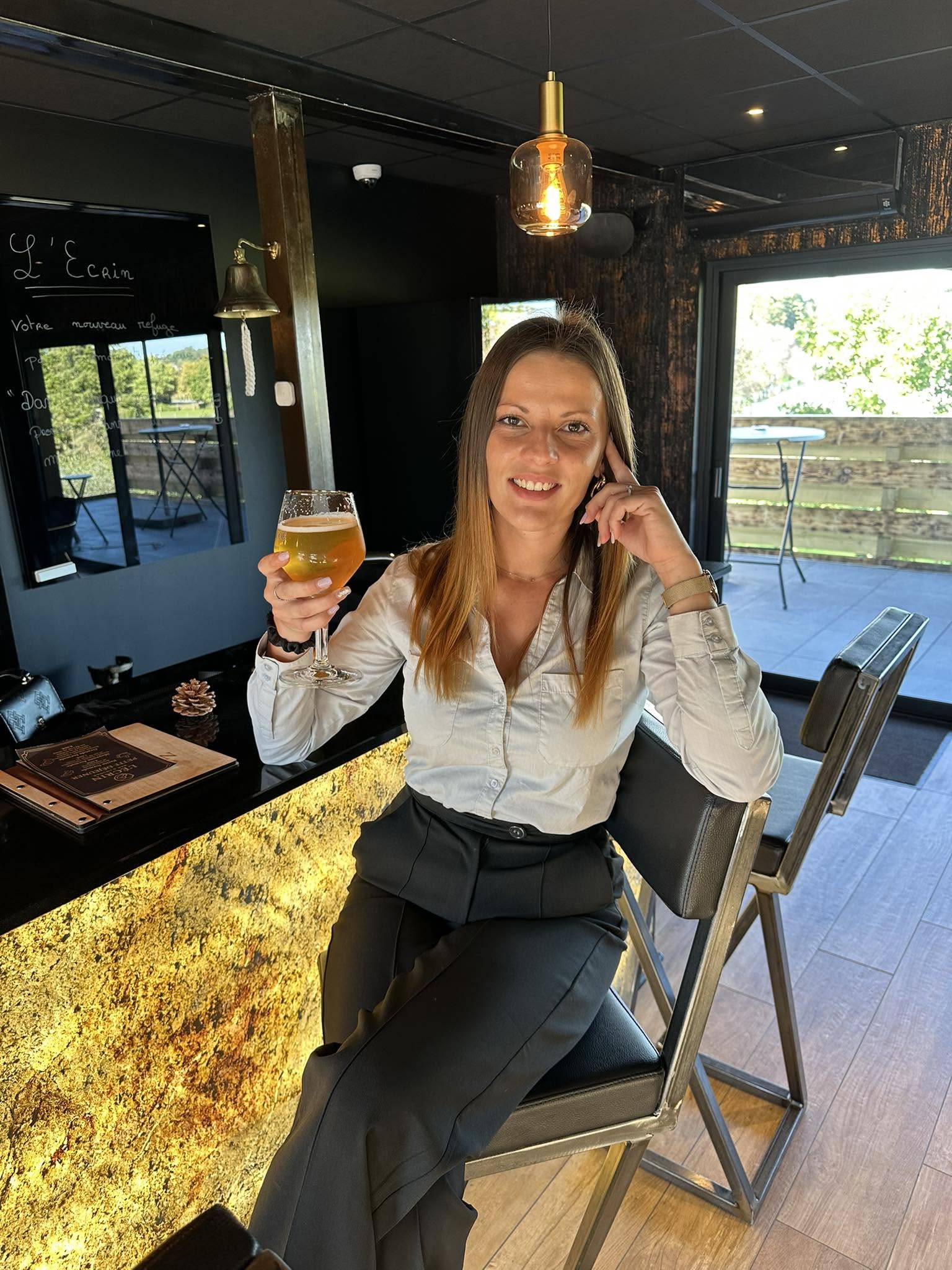 Laura au comptoir de L'Écrin, un verre à la main, la terrasse en arrière-plan